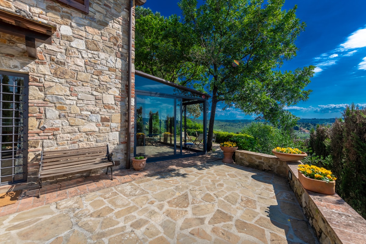 A stone terrace provides a spacious outdoor area, bordered by potted flowers and shaded by trees. Large glass panels offer views of the surrounding hills and valleys, allowing natural light to fill the space. A wooden bench invites relaxation in this serene setting.