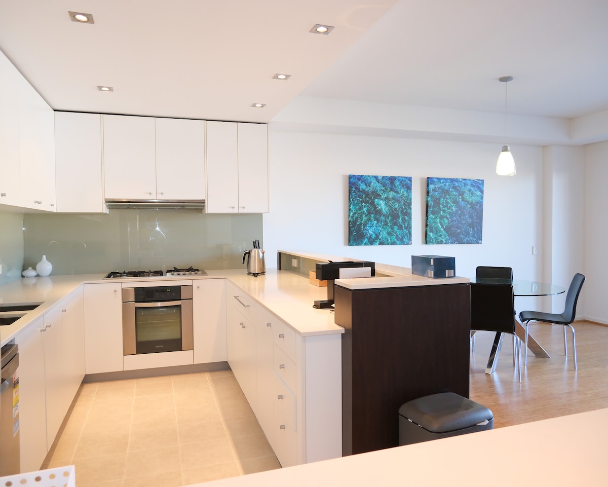 A modern kitchen features sleek white cabinetry and a spacious countertop. Stainless steel appliances, including an oven and kettle, are neatly arranged. A glass dining table with black chairs is positioned nearby, complemented by two large artworks on the wall.