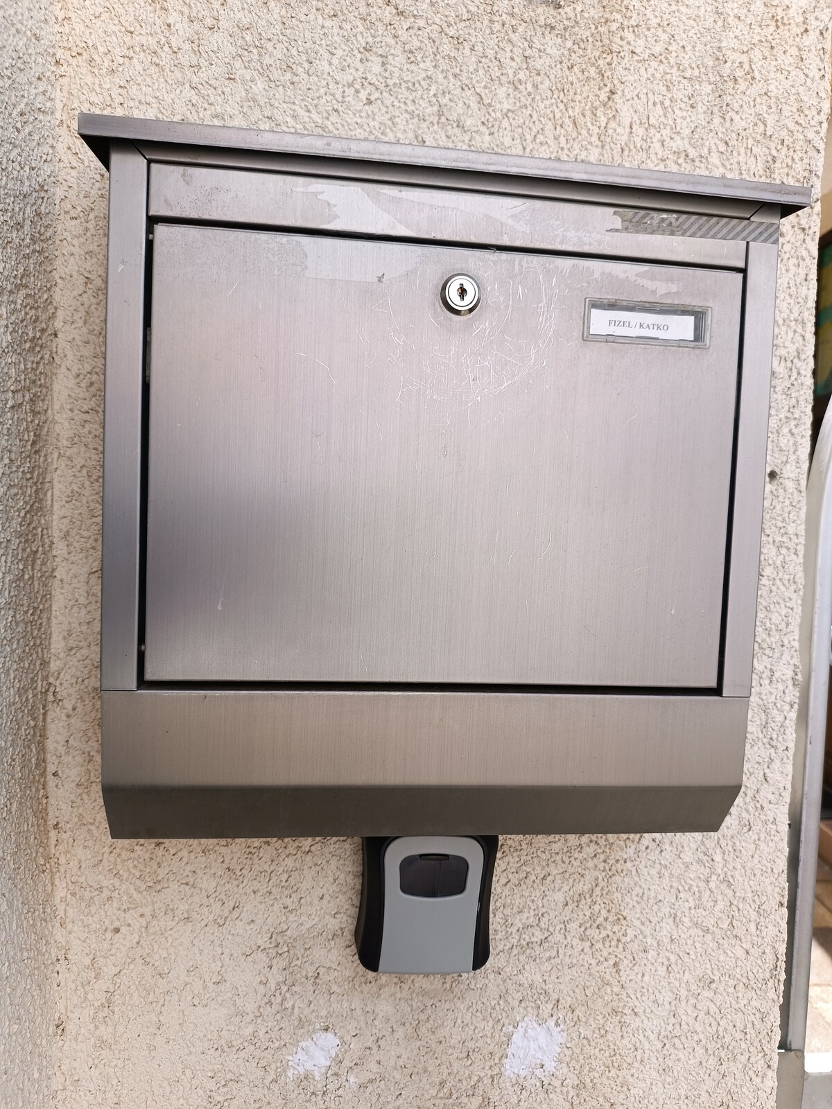 A metallic mailbox is mounted on a textured beige wall. The mailbox features a simple design with a secure locking mechanism and a label holder on the front.