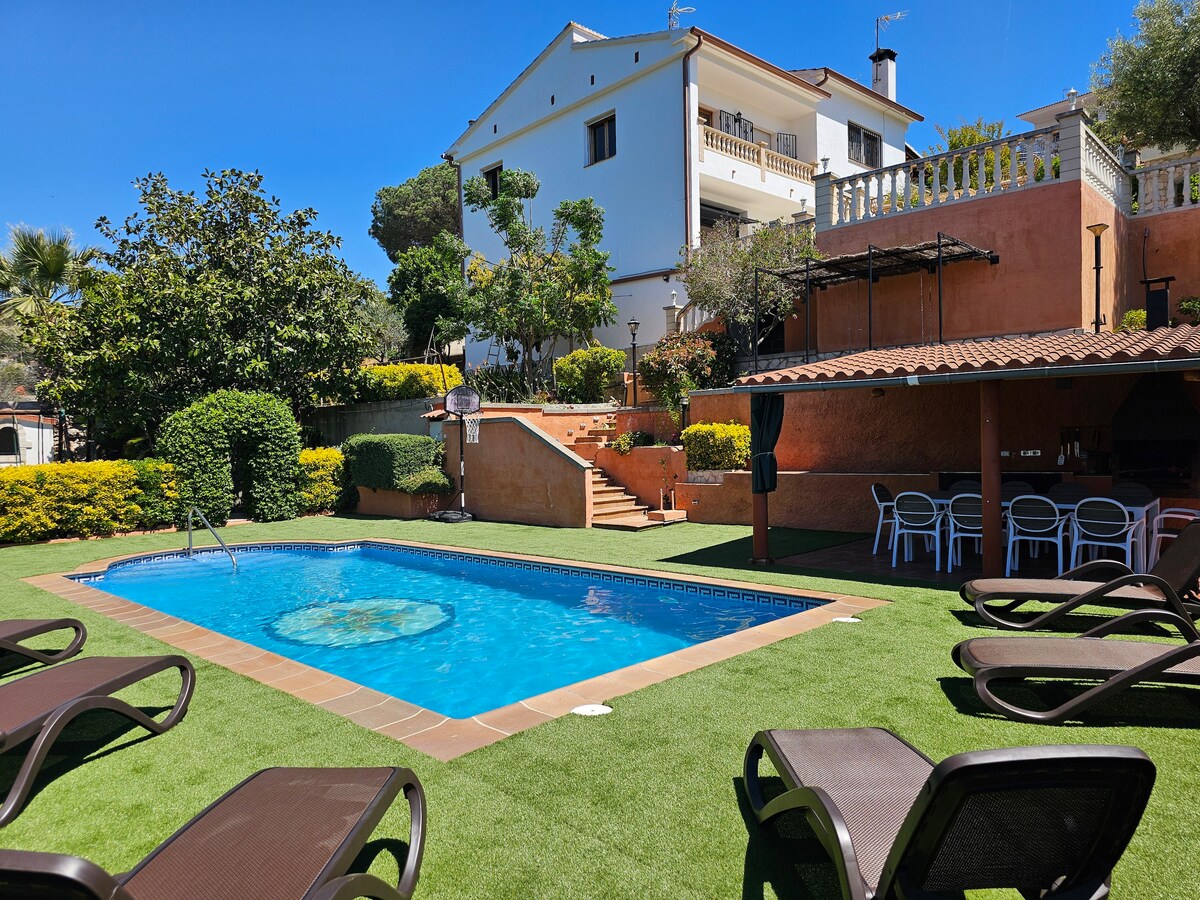 A private salt-water pool is surrounded by well-maintained green grass and sun loungers. The outdoor space features a shaded area with a dining setup and a barbecue. Lush plants and trees provide a tranquil atmosphere, complementing the modern villa in the background.