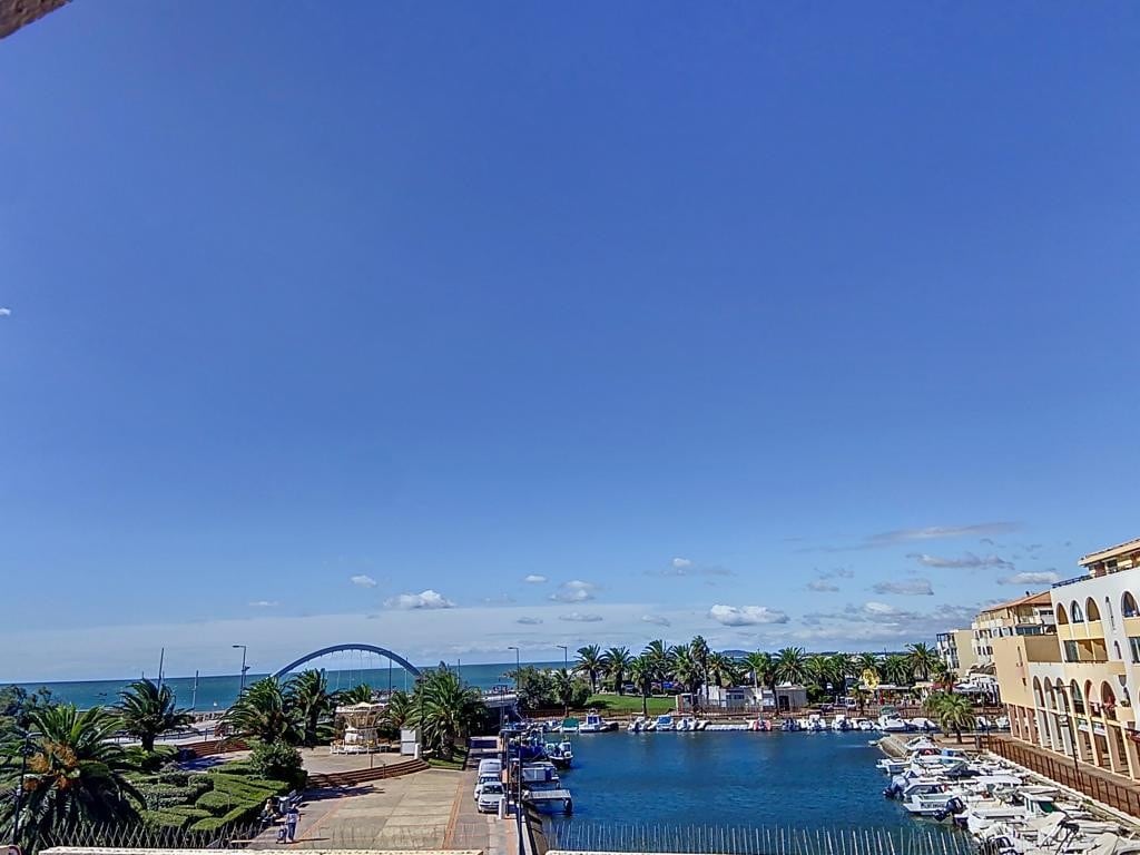 A serene coastal view showcases a marina filled with boats, bordered by lush palm trees. The ocean is visible beyond the marina, under a clear blue sky. A pedestrian area with stone pavement leads to the water, contributing to a relaxed atmosphere.