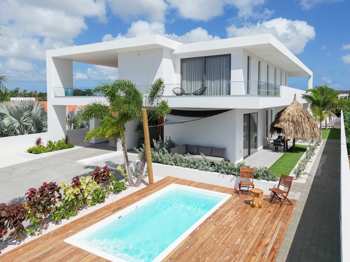 A modern two-story building features expansive glass windows and a balcony that overlooks a swimming pool. The outdoor area includes a wooden deck with lounge chairs and tropical landscaping, enhancing the pool's tranquil setting under a partly cloudy sky.