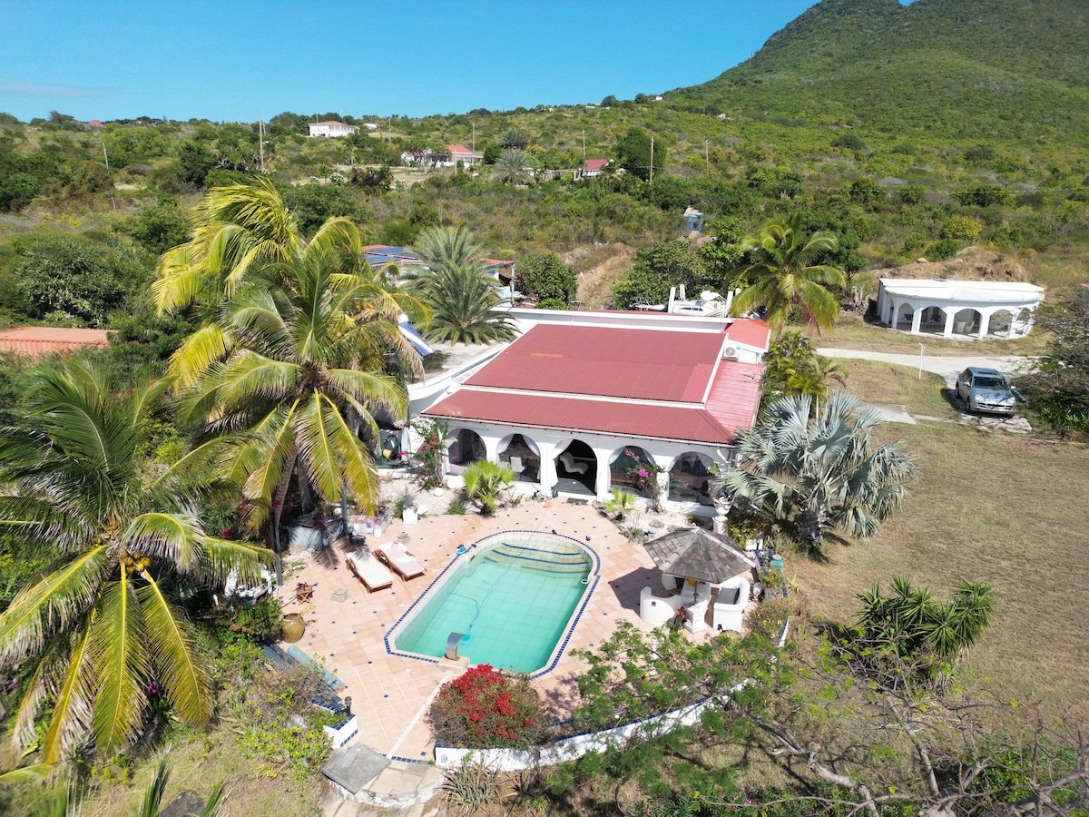 An aerial view reveals a sprawling property featuring a centrally located pool surrounded by tropical vegetation. Palm trees and colorful flowers enhance the outdoor space, while a red-roofed building complements the natural landscape, with distant hills providing a serene backdrop.