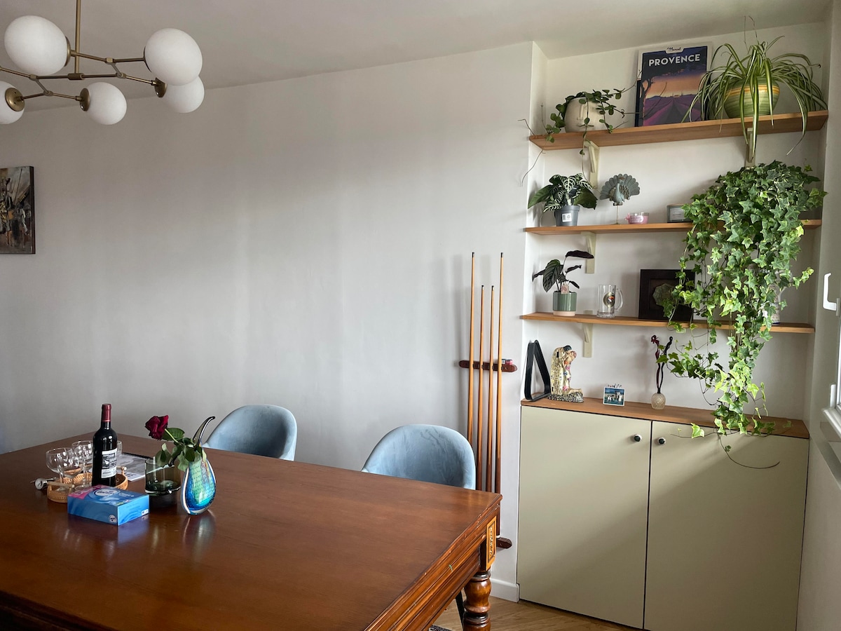A dining area is depicted with a wooden table and plush blue chairs. A shelf adorned with various plants and decorative items stands against the wall, complemented by a simple cabinet below. Natural light illuminates the space, enhancing the greenery.