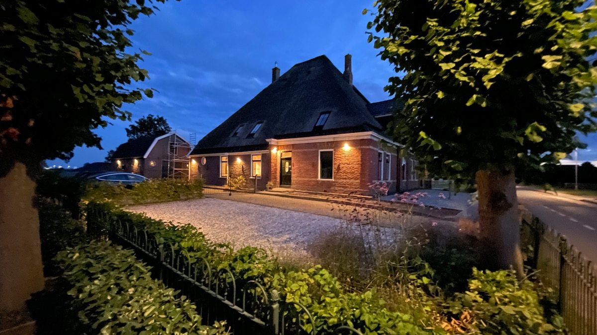 The exterior of a charming Dutch farmhouse is illuminated at dusk, showcasing its traditional thatched roof and red brick walls. Lush greenery and neatly trimmed hedges frame the gravel driveway, while soft lighting highlights the entrance and surrounding area.
