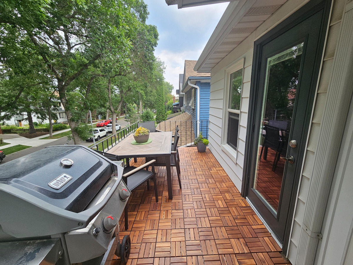 The outdoor balcony features a Weber barbeque and a sturdy table with four chairs. Potted plants add greenery to the space. The wooden flooring provides a warm appearance, while a railing ensures safety and views of the surrounding tree-lined neighborhood.