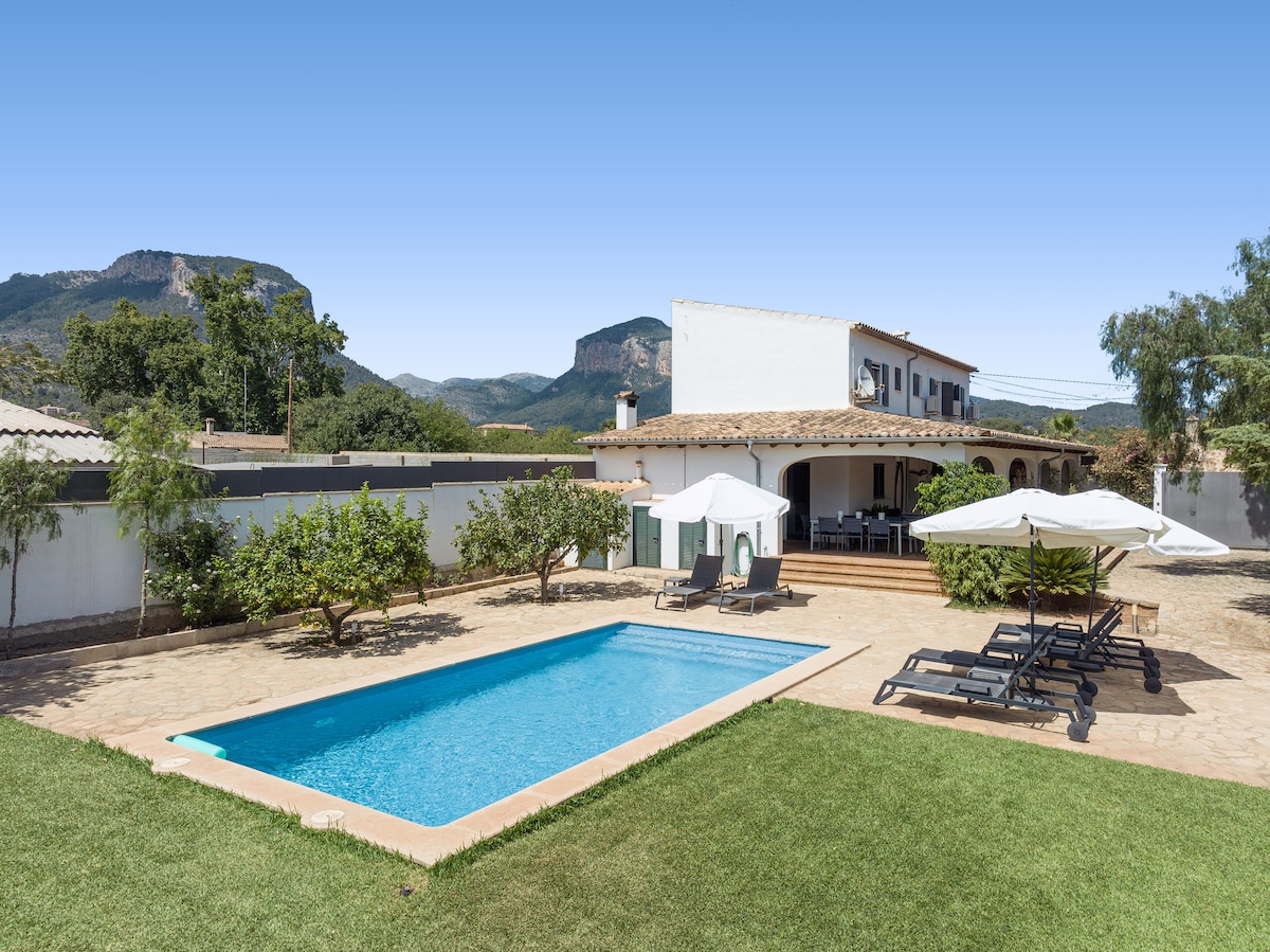 A spacious outdoor area features a salt-water pool surrounded by sun loungers and large umbrellas. Nearby, fruit trees provide shade, and a covered terrace offers seating options. The backdrop displays the picturesque Serra de Tramuntana mountains, completing this serene setting.