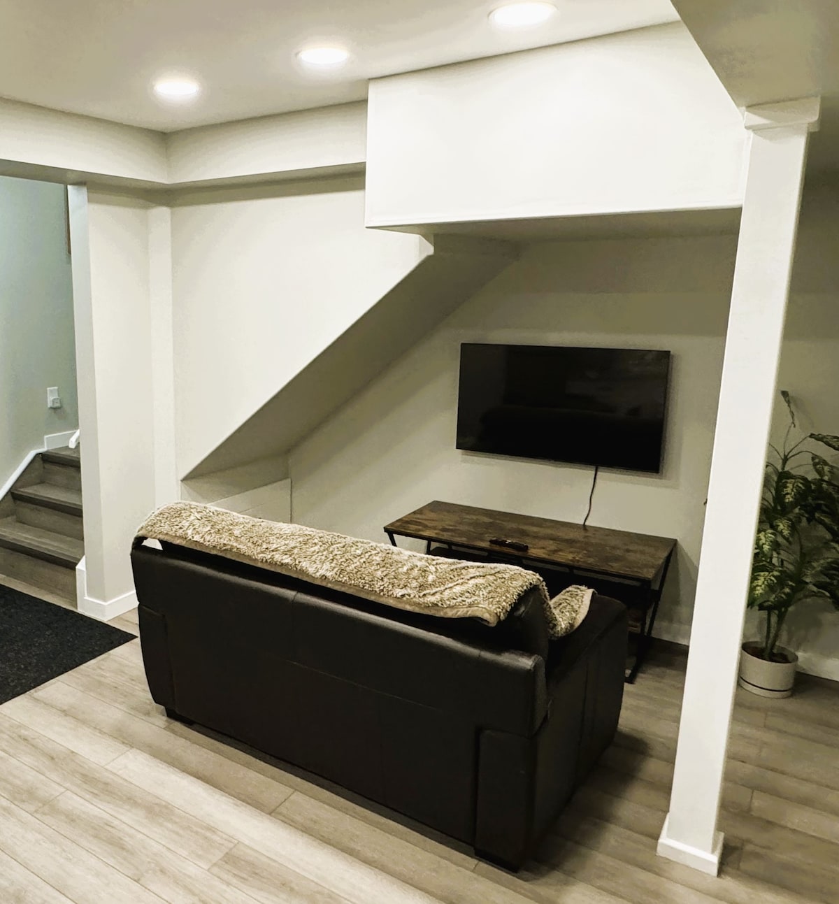 A cozy seating area features a dark leather couch draped with a light throw, positioned across from a wooden coffee table and a wall-mounted television. Soft recessed lighting enhances the minimalist design, with a potted plant adding a touch of greenery in the corner.