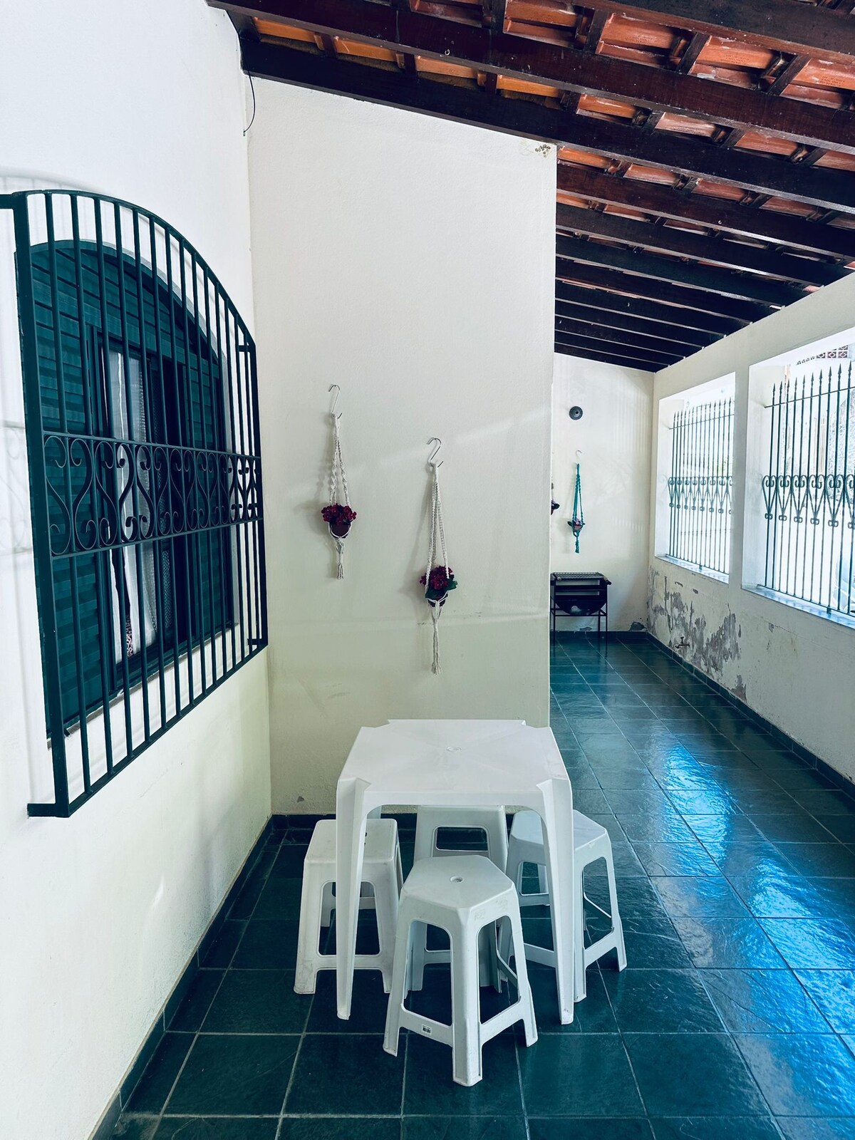 A narrow hallway is lined with green tiled flooring and features a light-colored wall. A small white table is accompanied by four matching stools. Decorative items hang from hooks, and natural light enters through tall, barred windows along one side.