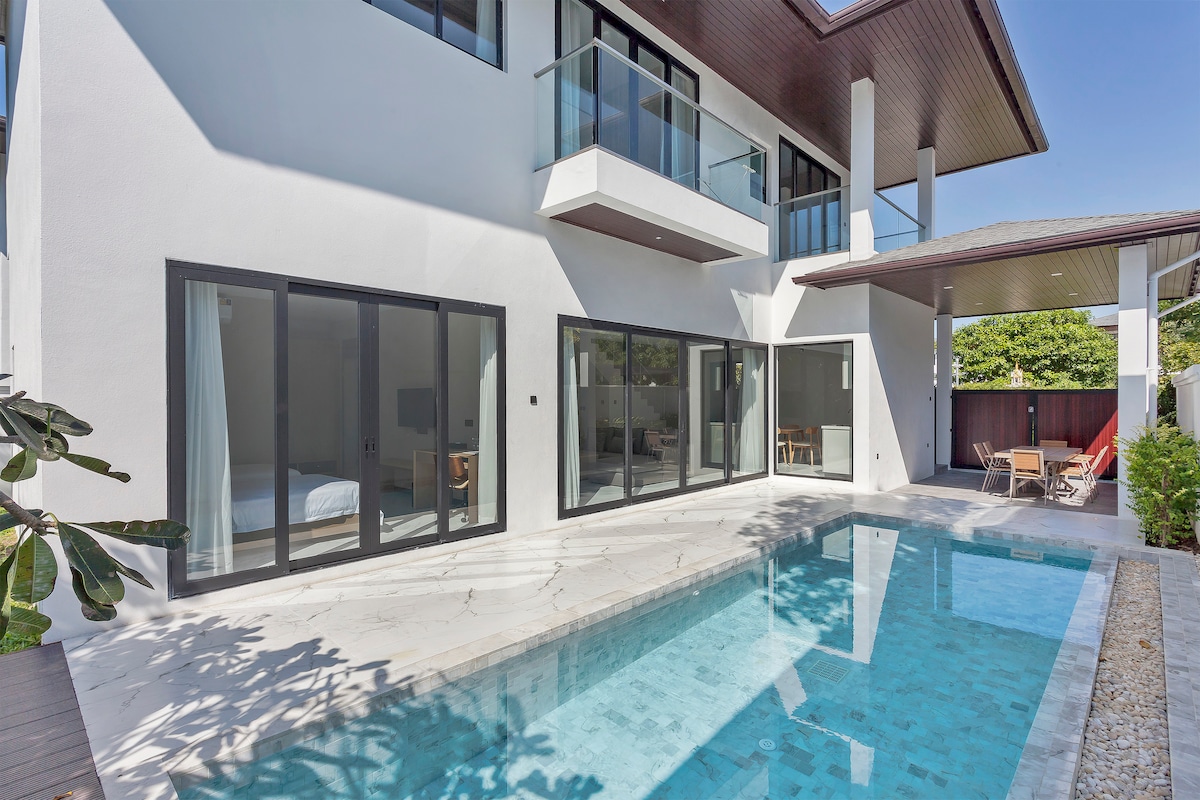 A modern villa exterior features large glass doors opening to a serene space. The private pool reflects light amidst marble flooring. Outdoor seating areas are visible near the water, surrounded by lush greenery and decorative plants.