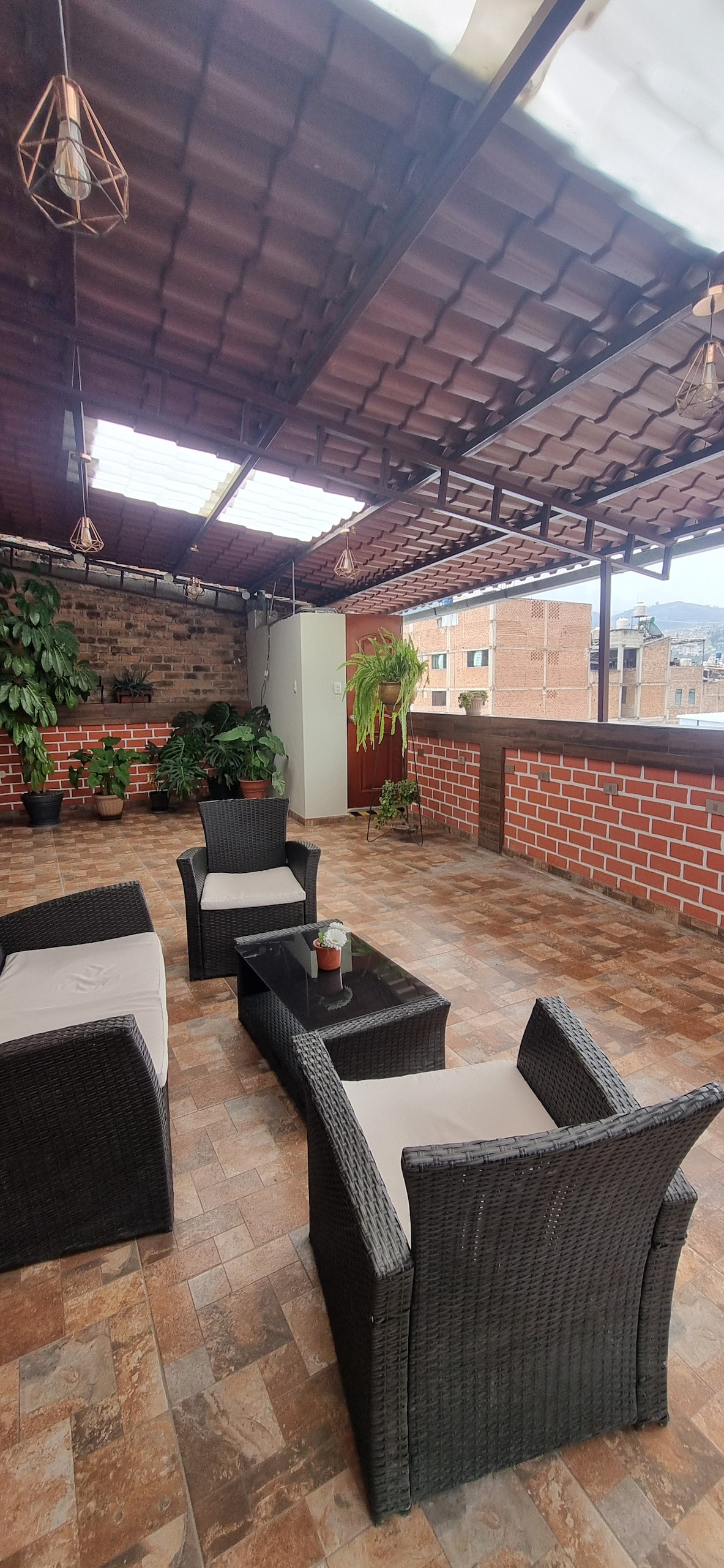 An outdoor terrace area is presented with dark wicker furniture, including four armchairs and a low table. Green plants are strategically placed against brick walls, enhancing the space. Natural light filters through a skylight, complementing the inviting ambiance of the setting.
