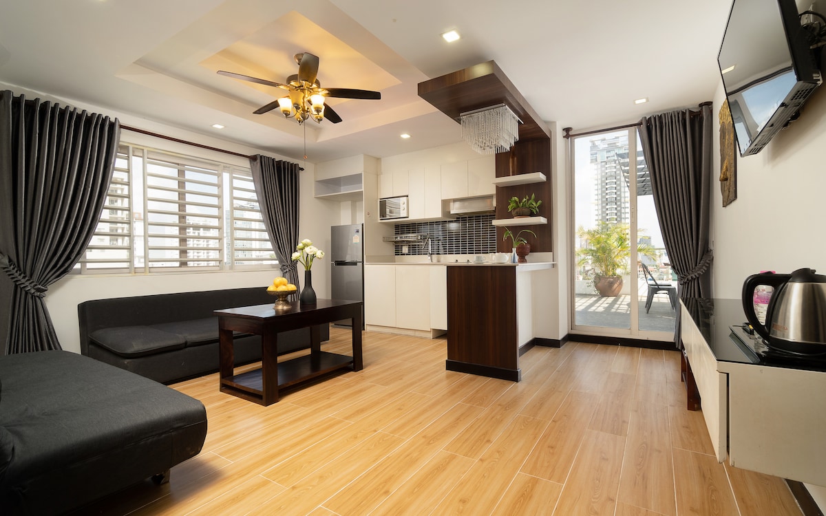 A modern living space features a black sofa and a wooden coffee table set amidst warm wooden flooring. A sleek kitchen area is visible, equipped with contemporary appliances. Large windows provide natural light, with curtains elegantly framing the view of a cityscape.