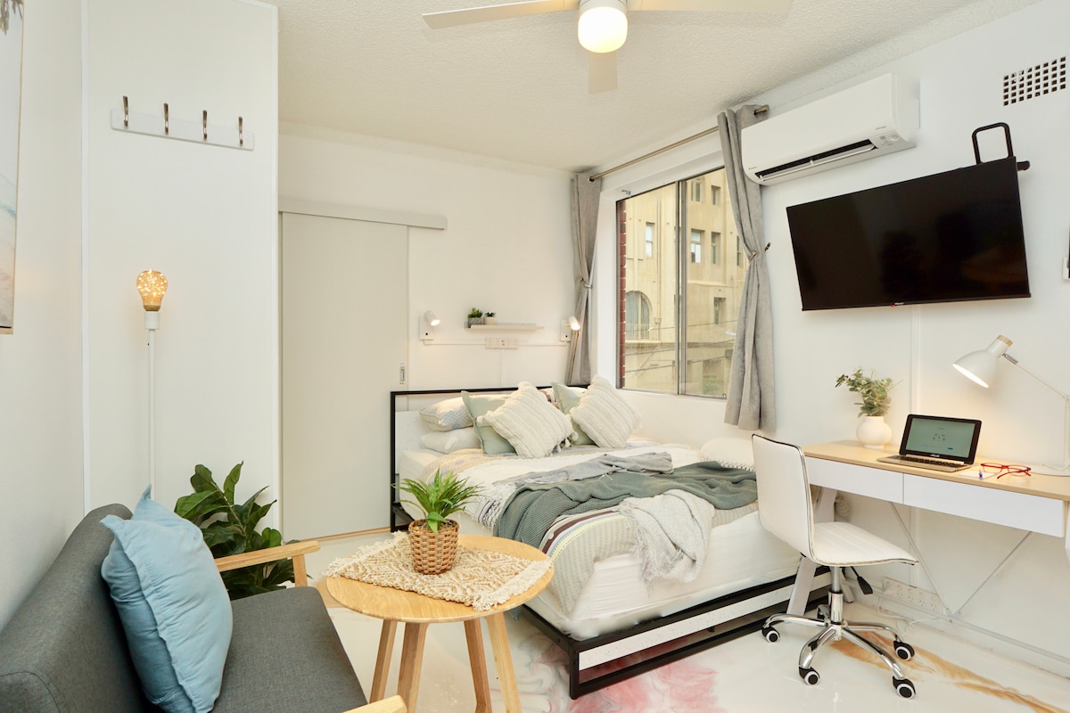 A modern studio space features a comfortable bed with layered linens, complemented by soft pillows. A cozy sofa with a throw pillow provides seating, while a small table holds a decorative plant. A wall-mounted TV and a workspace with a chair are also visible, along with large windows allowing natural light.