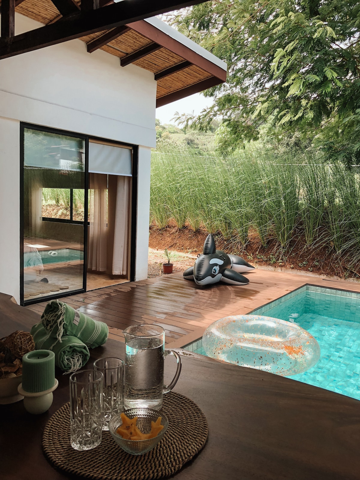 A view from the indoor space captures a serene pool area, complemented by a lounging inflatable. Natural light flows into the room, highlighting the wooden deck and surrounding greenery. Glassware and decor sit on a wooden table, offering a sense of relaxation and comfort.