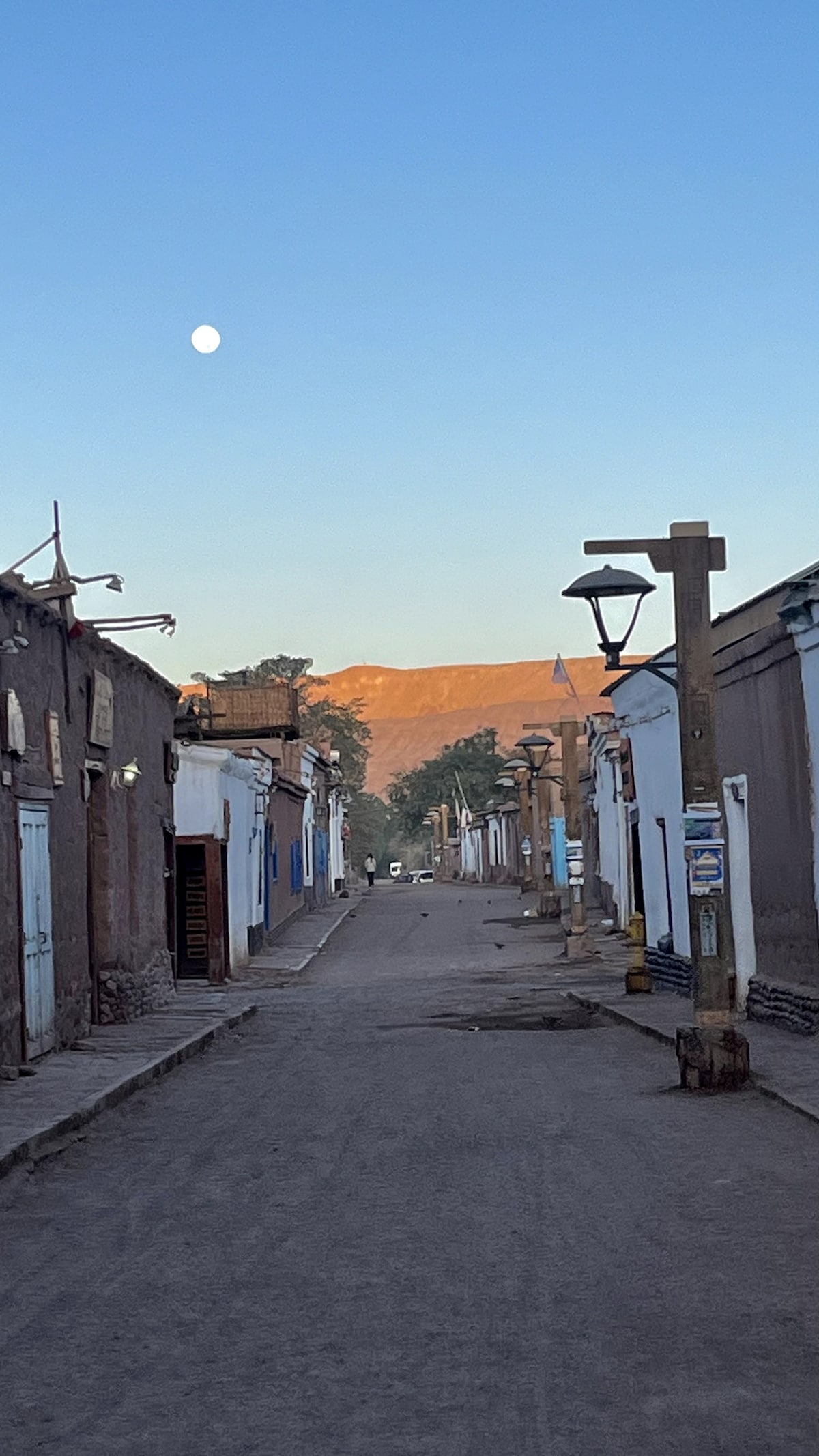 A quiet dirt street lined with rustic adobe buildings is seen under a clear blue sky. A gentle moon is visible, and soft sunlight lights up the distant mountains. Street lamps are strategically placed along the pathway, enhancing the tranquil ambiance.