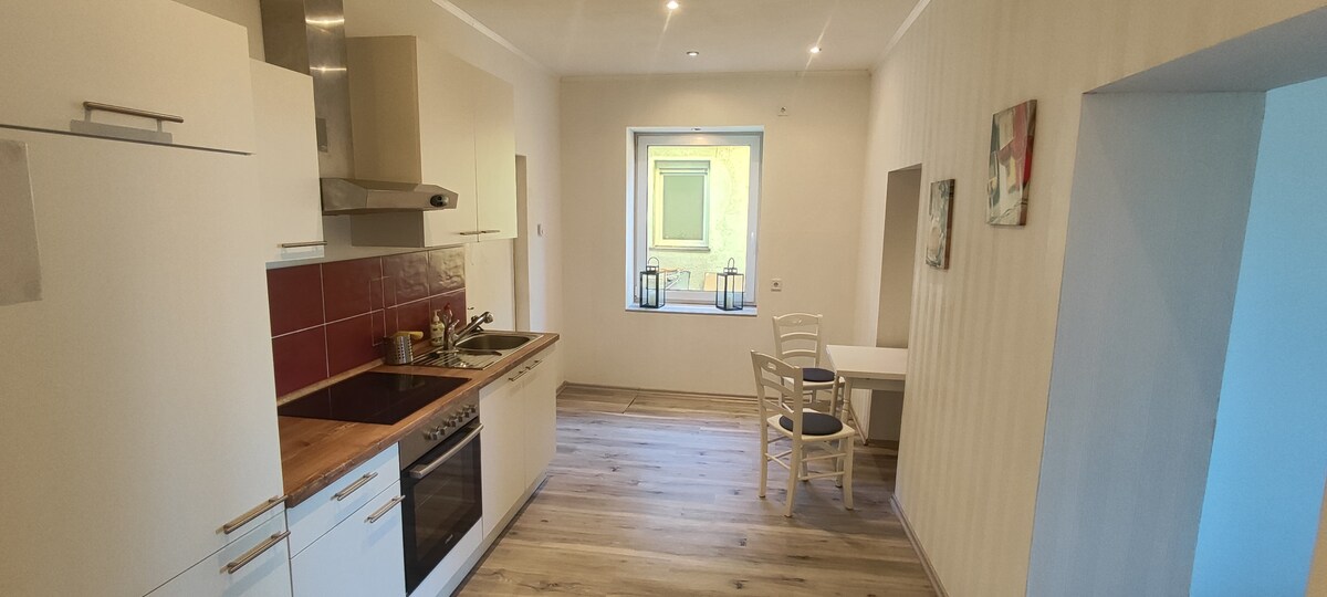 The kitchen features modern cabinetry with a wooden countertop and integrated appliances. A small dining table with two chairs is present, positioned near the window, which offers natural light. Light-colored walls and wooden flooring enhance the spacious feel of the area.