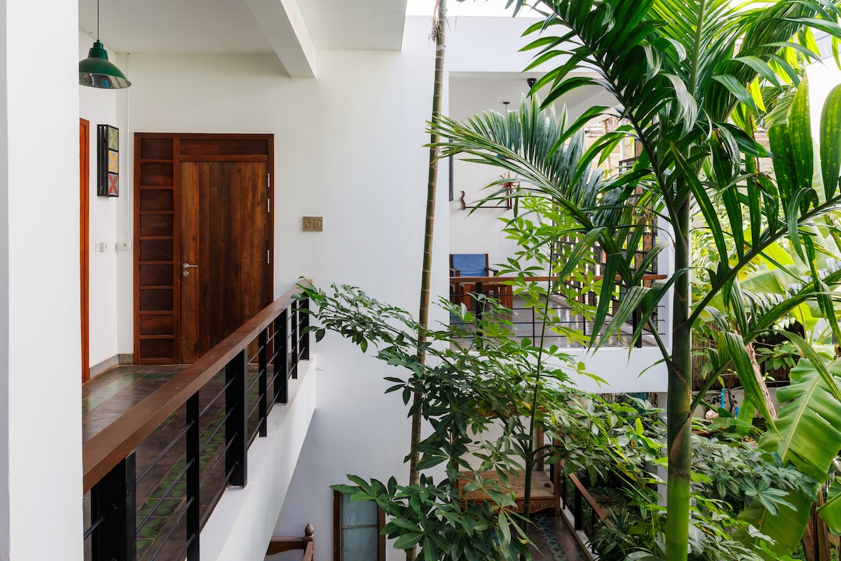 A serene corridor is seen, surrounded by lush greenery and tropical plants. Natural light floods the space, highlighting a wooden door and balcony railing. Comfortable seating areas are visible on the levels above, creating an inviting atmosphere amidst the greenery.