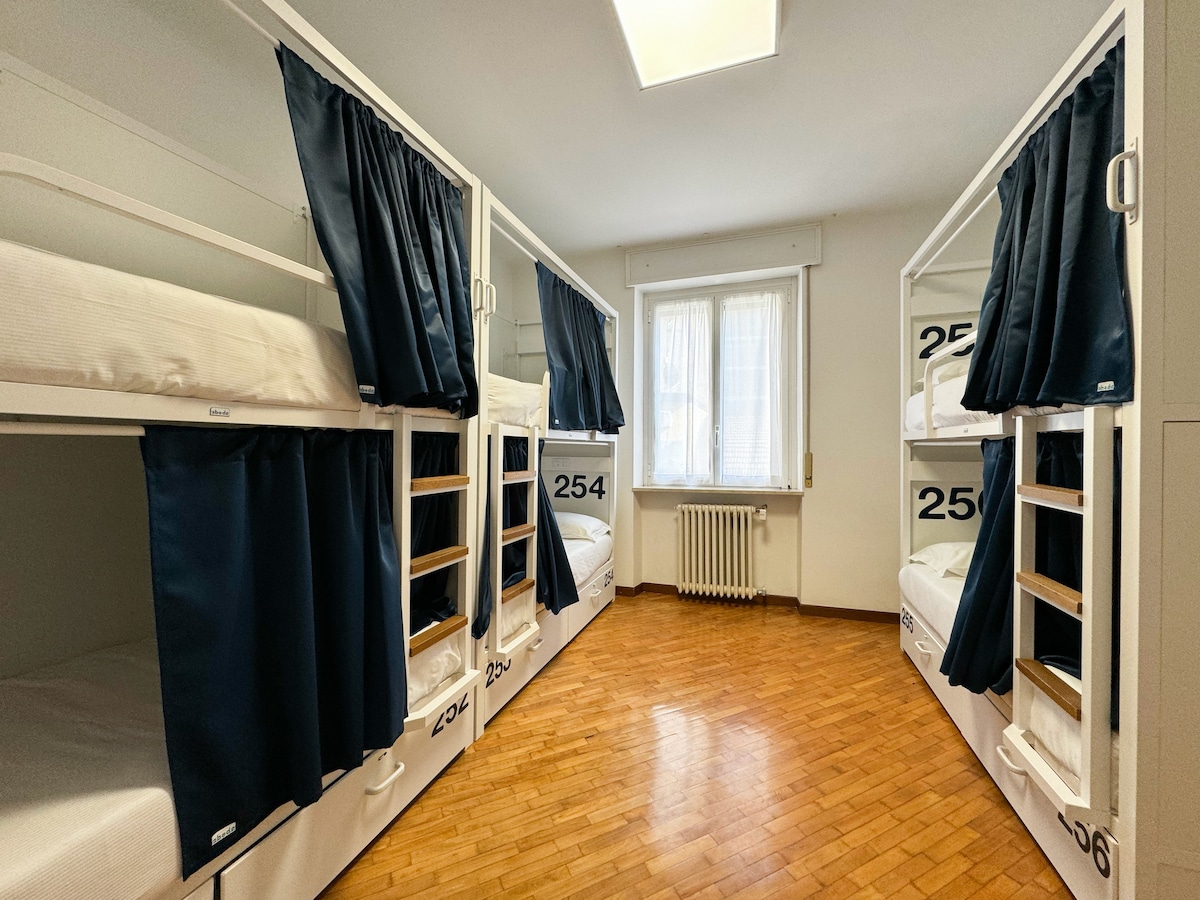 The dormitory features three bunk beds, accommodating six guests. Each bed is enclosed with a curtain for privacy. A bright window allows natural light to fill the space, highlighting the warm wooden flooring and simple, functional design.