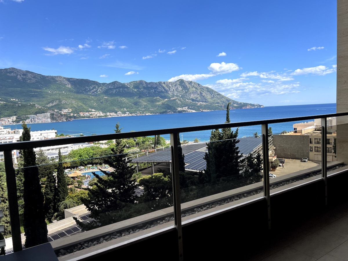 A balcony view captures the expansive coastline, framed by mountains in the background. A clear blue sky complements the tranquil sea, while lush greenery is visible below. The glass railing provides an unobstructed perspective of the vibrant landscape.