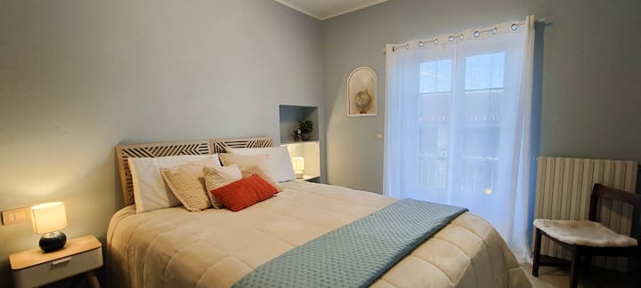 The room is furnished with a large and comfortable double bed, complete with quality mattress and soft linens. Two nightstands with lamps offer soft lighting, while the window with light curtains ensures privacy. 