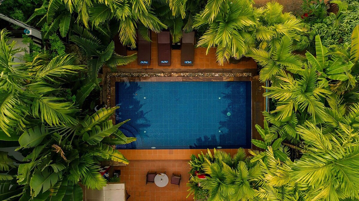 An aerial view captures a serene swimming pool surrounded by lush tropical greenery. Comfortable lounge chairs are positioned on the pool's deck, and a small round table with chairs is visible nearby, offering a perfect spot to relax in the vibrant environment.
