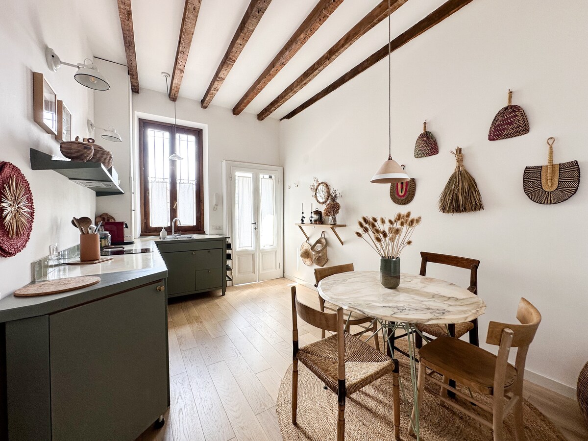 The kitchen area features high wooden ceilings with exposed beams and a circular dining table surrounded by wooden chairs. Decorative wall elements add texture, while a doorway opens to the outside. The space is well-lit by natural light, creating a welcoming atmosphere.