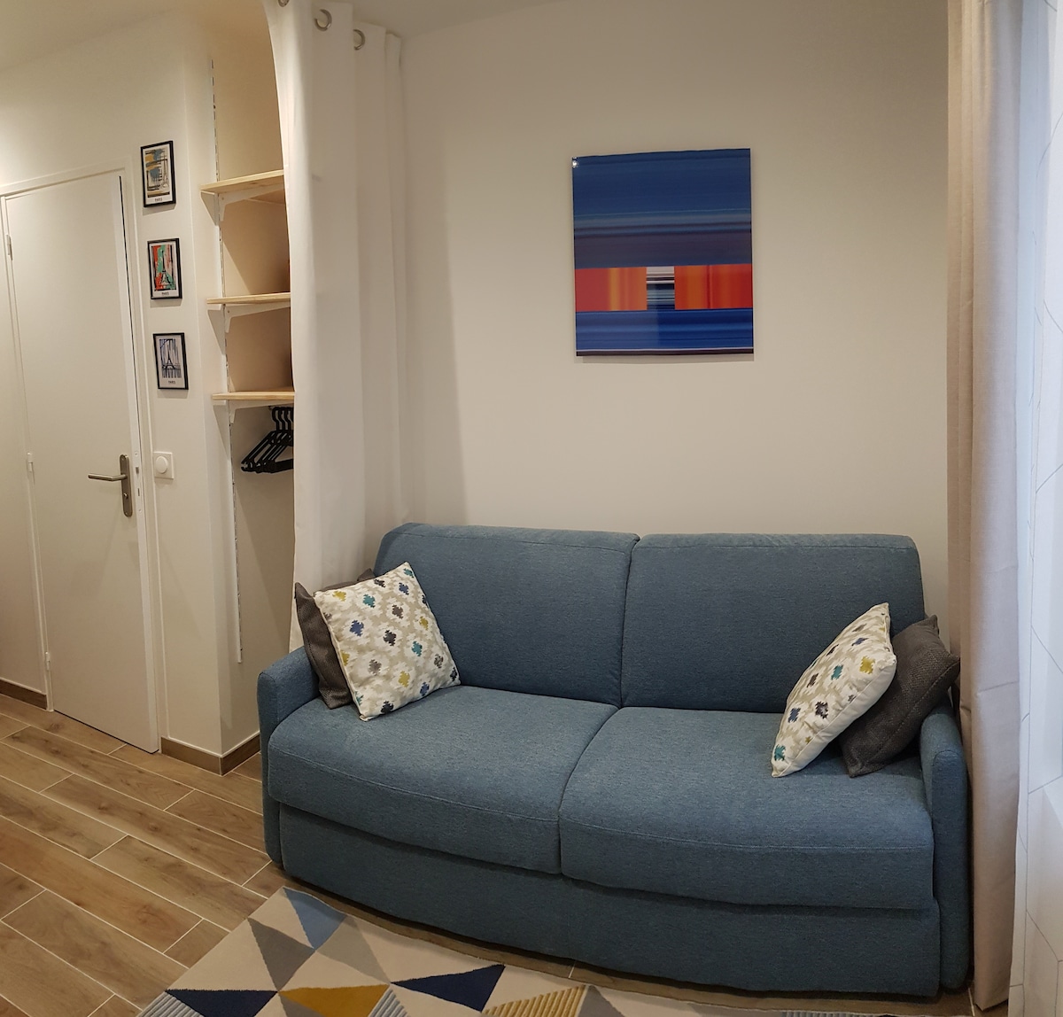 A cozy seating area is highlighted by a spacious blue sofa adorned with decorative pillows. Above the sofa, a modern piece of artwork enhances the wall, while a nearby shelving unit provides additional storage. Natural light flows in through a window partially covered by sheer curtains.
