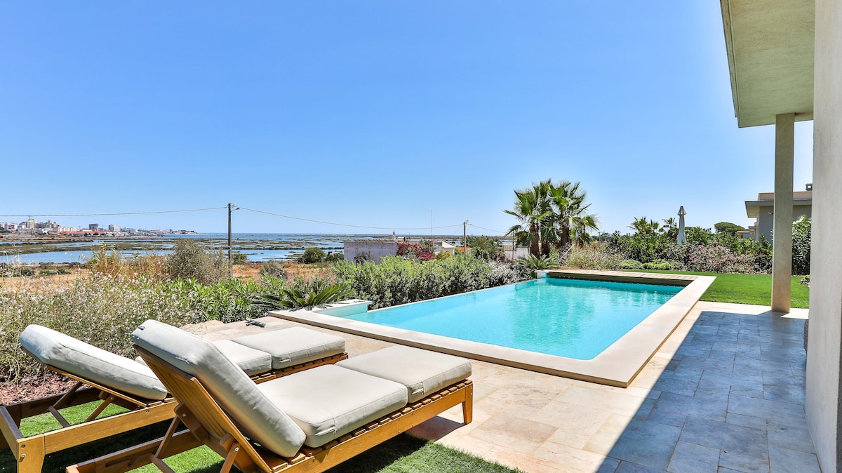 A serene outdoor space features a private heated swimming pool, surrounded by landscaped gardens. Comfortable lounge chairs lie next to the pool, offering a view of the Ria Formosa and distant Faro city. Clear blue skies enhance the peacefulness of the setting.