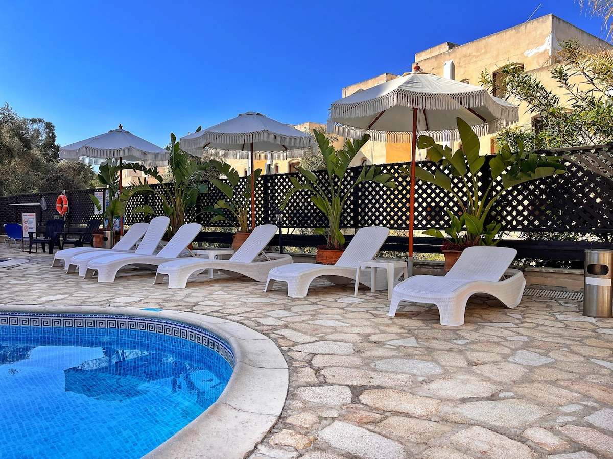 A communal swimming pool area features several white sun loungers arranged around the water's edge. Large umbrellas provide shade, while tropical plants are placed in terracotta pots along the perimeter. The pool is bordered by a patterned tile edge, enhancing the serene ambiance.