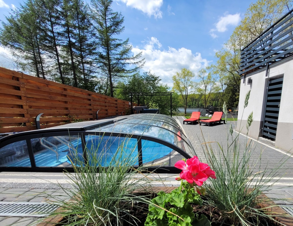 An enclosed swimming pool is visible, surrounded by greenery and a wooden fence. Bright red lounge chairs are positioned nearby, offering a place to relax. A vibrant pink flower adds a touch of color to the foreground, complementing the serene outdoor atmosphere.