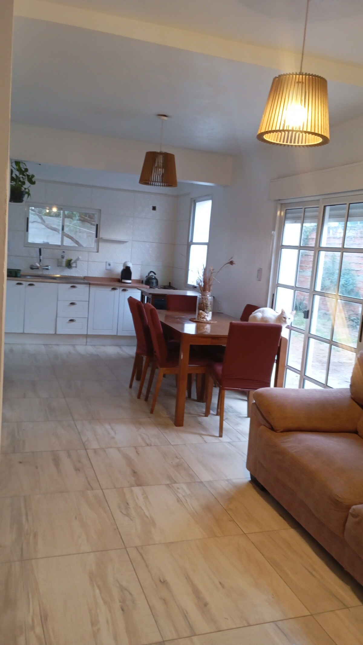 A spacious dining area is equipped with a round wooden table surrounded by four chairs. Natural light enters through large glass doors, creating a bright atmosphere. A comfortable seating area with a soft sofa is positioned nearby, enhancing the open layout of the living space.