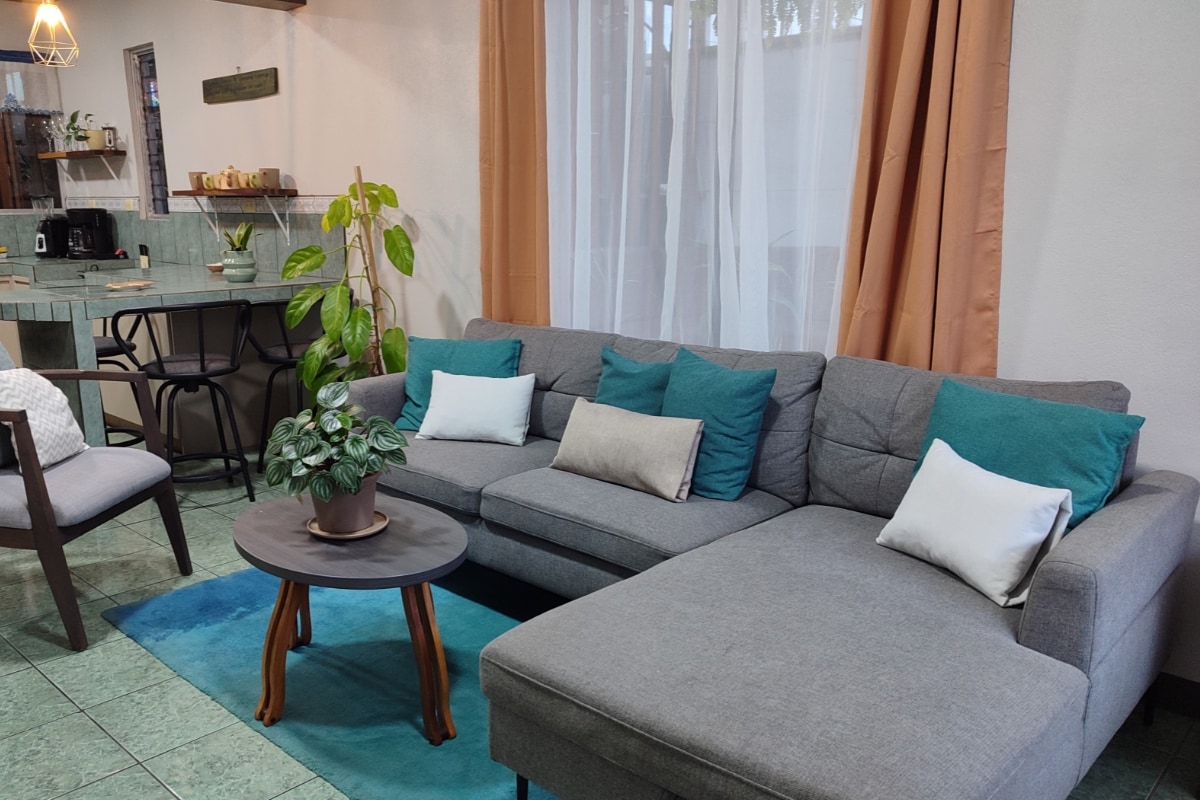 A contemporary living area features a gray sectional sofa adorned with cushions in soft hues. A round wooden coffee table with a small potted plant sits in the center. Light filters through sheer curtains, complementing the green tiled flooring and the kitchen area visible in the background.
