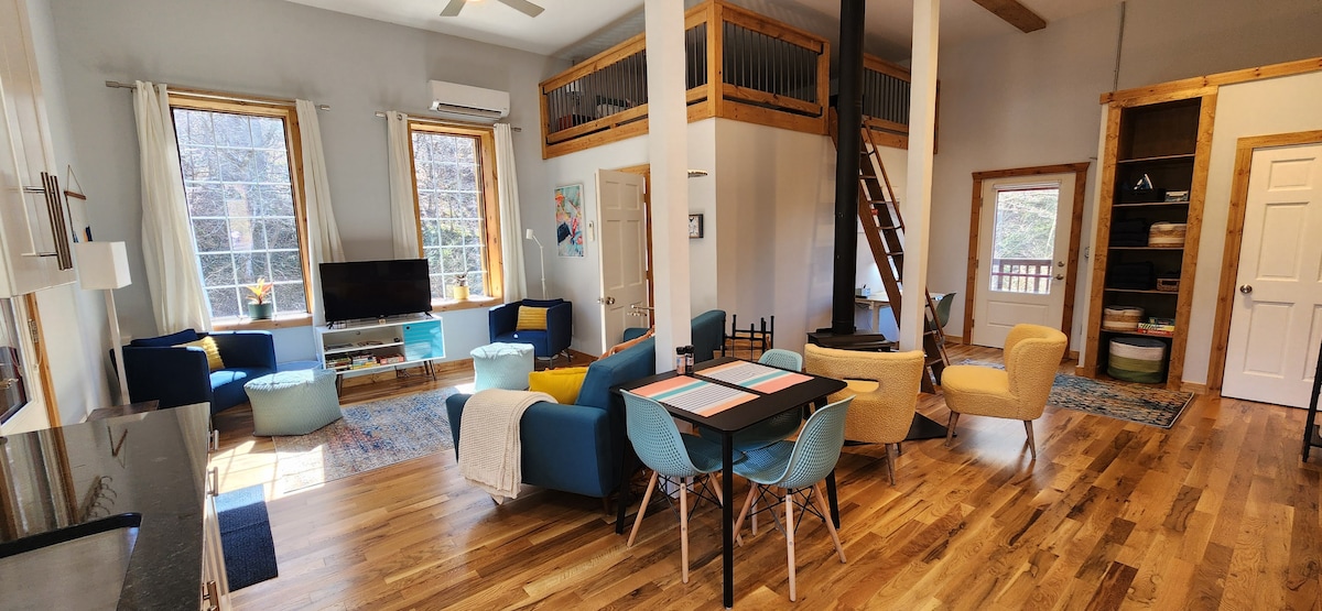 A spacious living area features large windows allowing ample natural light. Comfortable seating arrangements are positioned around a central dining table. A staircase leads to an upper loft, while modern accents and hardwood flooring enhance the space.