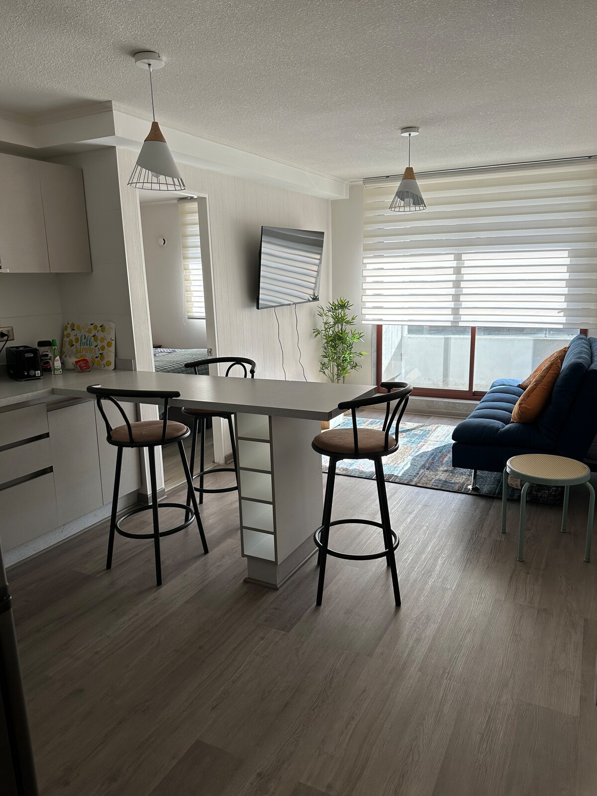 An open kitchen and living area is visible, featuring a modern breakfast bar with two high stools. A comfortable blue sofa is positioned near the window, complemented by light filtering through sheer blinds. A flat-screen TV is mounted on the wall, enhancing the space.