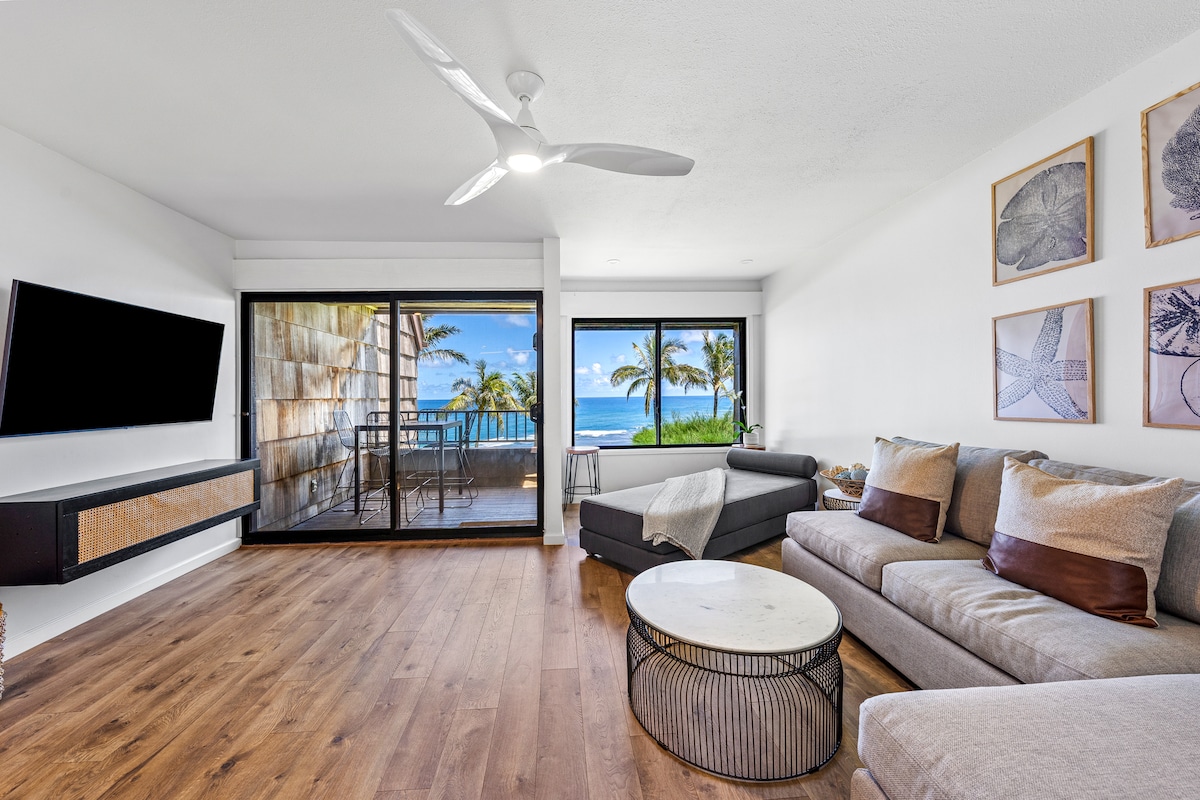 A spacious living area features a large window and sliding glass doors, providing a view of the tranquil Pacific Ocean and palm trees beyond. Furnished with a comfortable chaise sofa, a coffee table, and a wall-mounted TV, the room offers a modern and inviting space for relaxation.