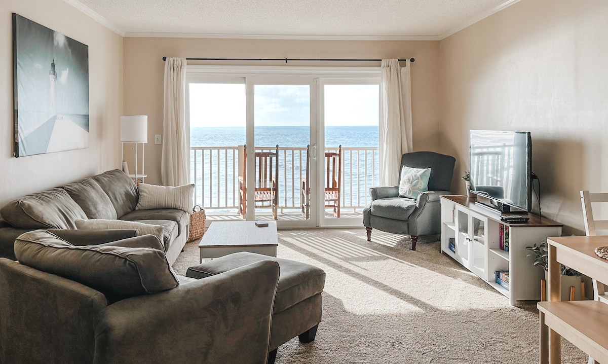 A bright living area features a comfortable sectional sofa and armchair, with a coffee table positioned centrally. Large sliding doors open to an ocean view, with two wooden rocking chairs on the balcony. The room is well-lit by natural light streaming through the windows.