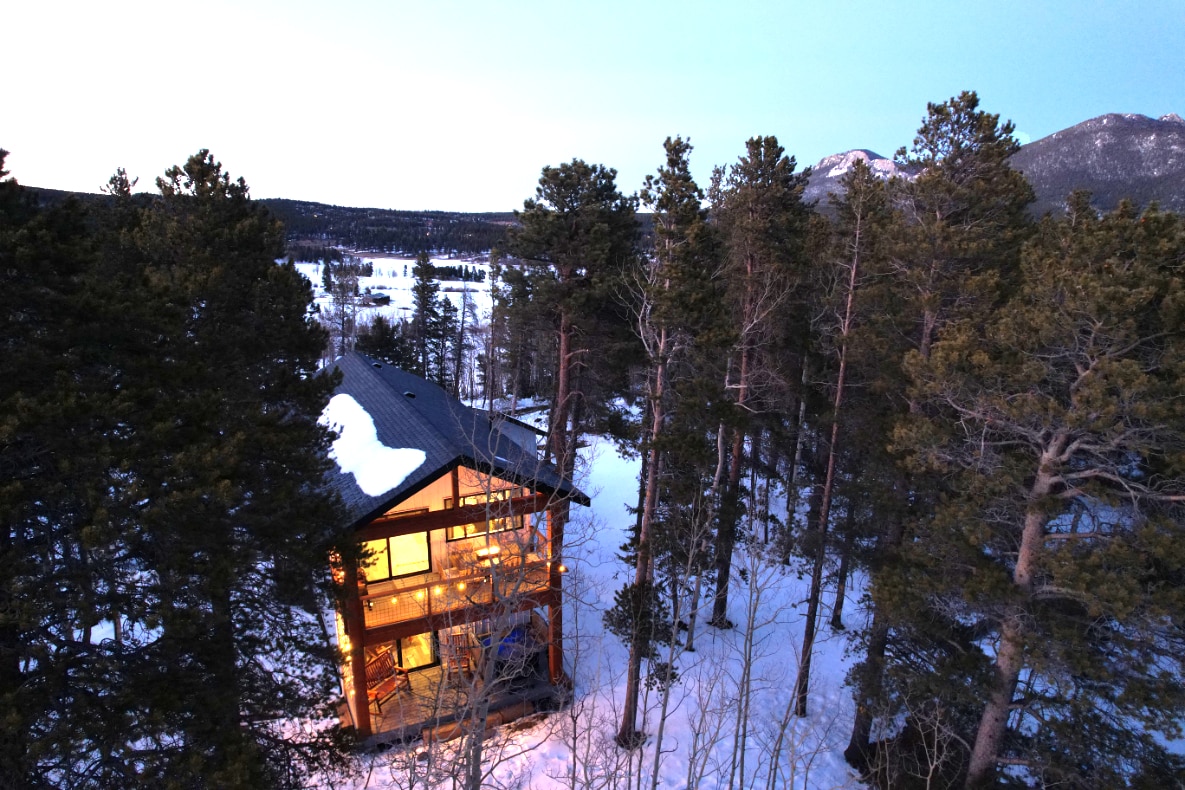 The exterior of the farmhouse is framed by tall pine trees, with a blanket of snow covering the ground. The structure is illuminated, creating a warm glow against the cool tones of twilight. Mountain peaks are visible in the distance, providing a serene backdrop.