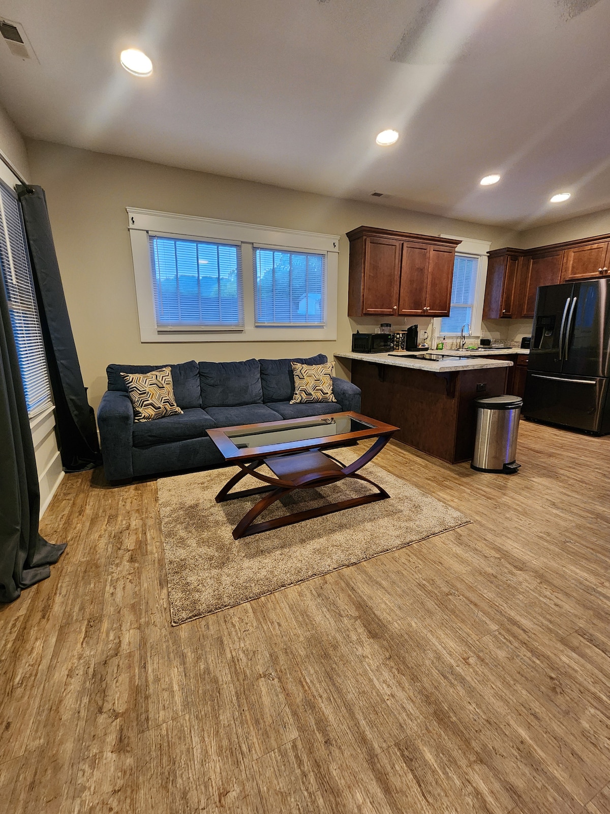 A living area features a comfortable blue sofa adorned with decorative pillows, positioned near a modern coffee table. Light from three windows illuminates the space, complemented by wooden cabinetry and a stainless-steel refrigerator. A rug adds texture to the hardwood flooring.