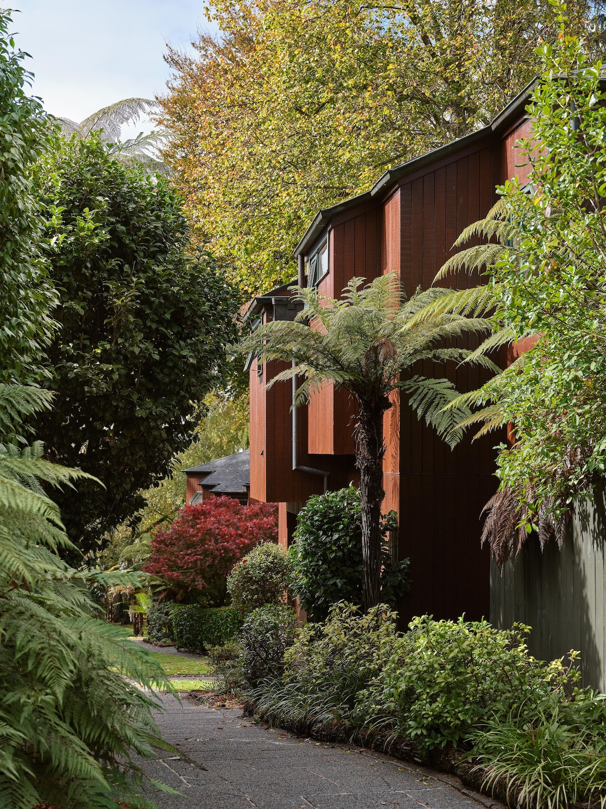 The exterior of Chalet #3 is framed by lush greenery and mature trees. The building features a rich, earthy hue and is two stories high, surrounded by well-maintained shrubs and plants that add to the natural setting. A pathway leads guests to the entrance.