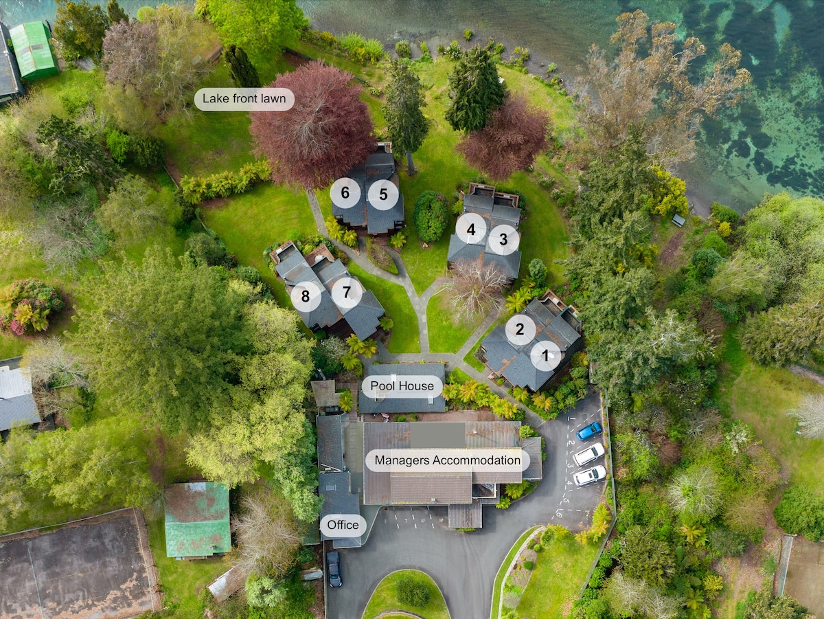 An aerial view of the property showcases the layout of the chalets numbered 1 to 8, surrounding a central pool house. Lush greenery, including trees and landscaped gardens, is visible, along with the lakefront lawn near the water's edge.