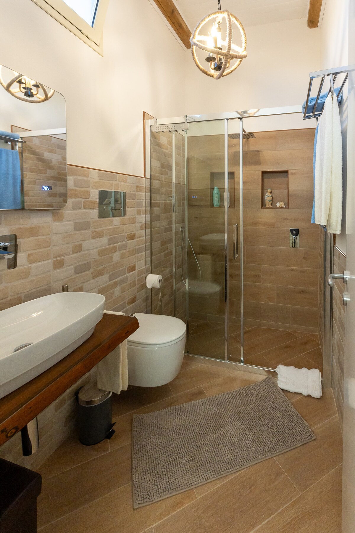 A modern bathroom features earthy tile walls and wood accents. A glass shower enclosure is positioned beside a contemporary toilet. A vessel sink rests on a wooden countertop, complemented by a soft bath mat on the floor. Natural light enters through a small window.