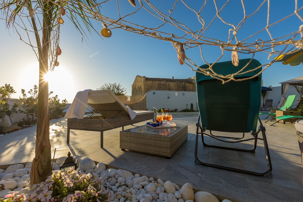 A sunlit terrace features a rustic seating area with a low table surrounded by lounge chairs. Decorated with seashells, the space is bordered by decorative stones and lush greenery. The silhouette of a historic building is visible in the background under a clear blue sky.