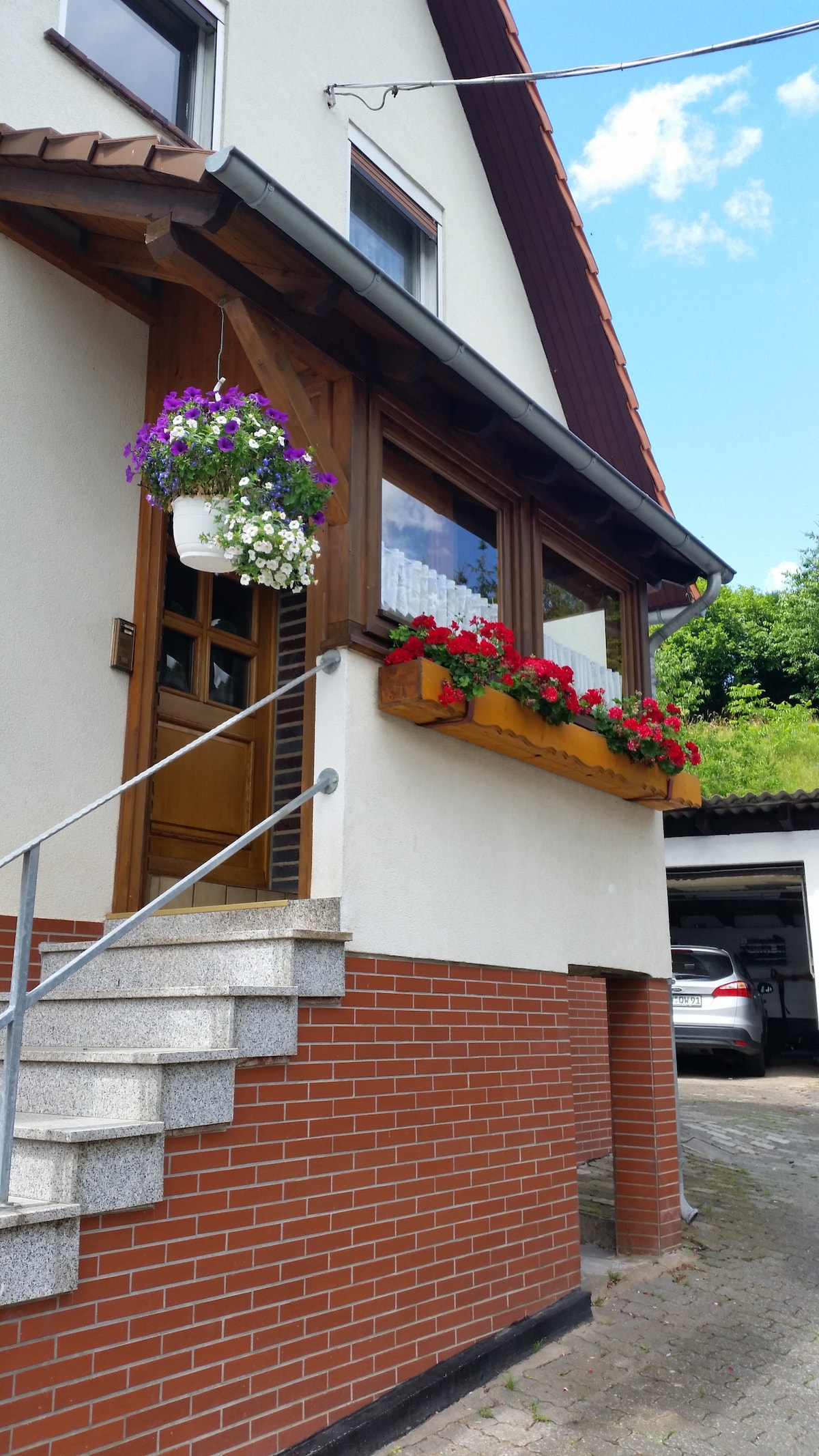 A charming entrance showcases a wooden door framed by vibrant flower boxes filled with colorful blooms. A set of steps leads up to the door, complementing the home’s exterior, with a driveway visible to the side.