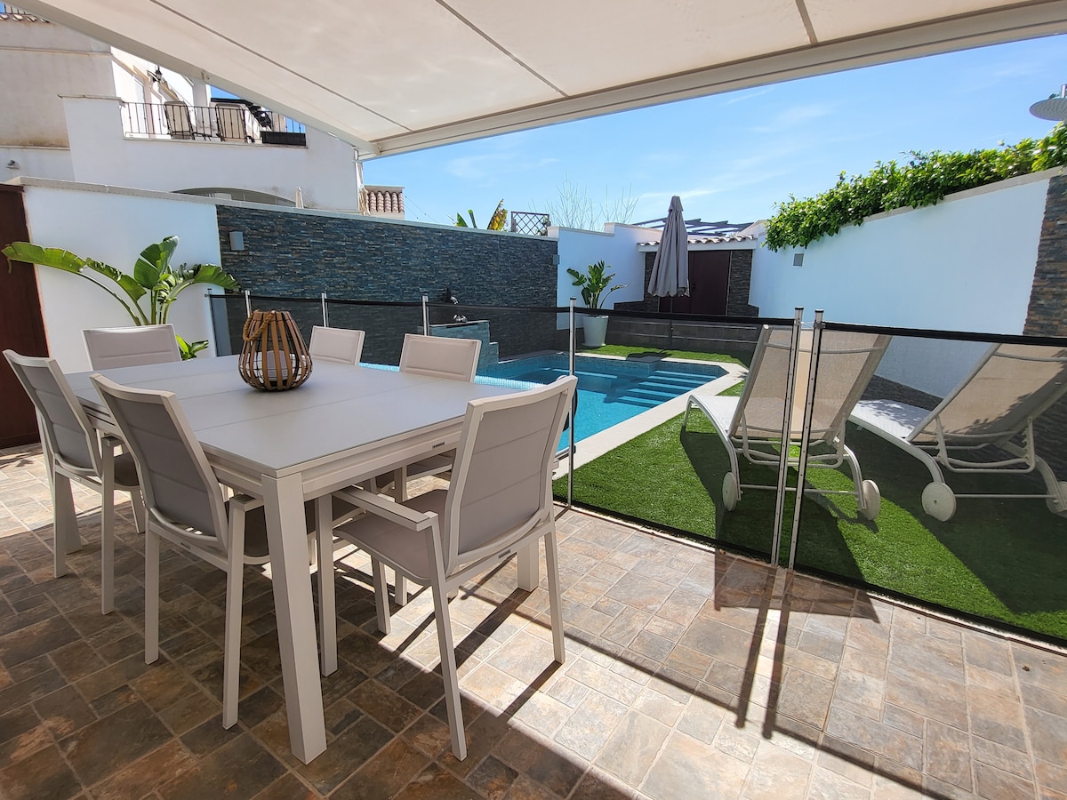 An outdoor dining area is presented with a large table and gray chairs, overlooking a well-maintained pool area. Sun loungers are positioned beside the pool, with lush greenery creating a relaxed atmosphere. A protective glass barrier enhances safety and visibility.