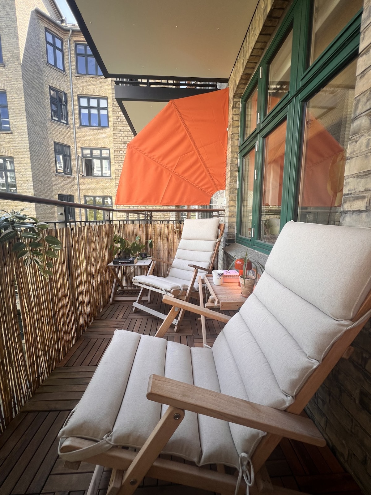 A balcony area features two comfortable seating chairs with soft cushions, positioned to enjoy the outdoors. A vibrant orange umbrella provides shade, while wooden flooring adds warmth. Potted plants enhance the greenery, and a small table is visible, suggesting a relaxing space.