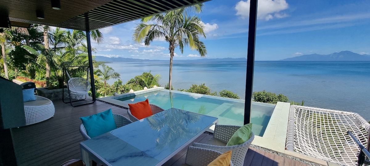 An outdoor lounge area features a modern white table surrounded by colorful cushions. A serene infinity pool blends seamlessly with the ocean view, while palm trees sway gently in the breeze, framing a tranquil atmosphere and expansive sea horizon under a clear blue sky.