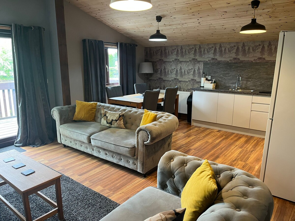 An open-plan living area is featured, with a plush gray sofa adorned with yellow cushions. A dining table with dark chairs is positioned near a modern kitchen, characterized by white cabinetry and a textured stone backsplash. Natural light is let in through large windows, enhancing the inviting space.