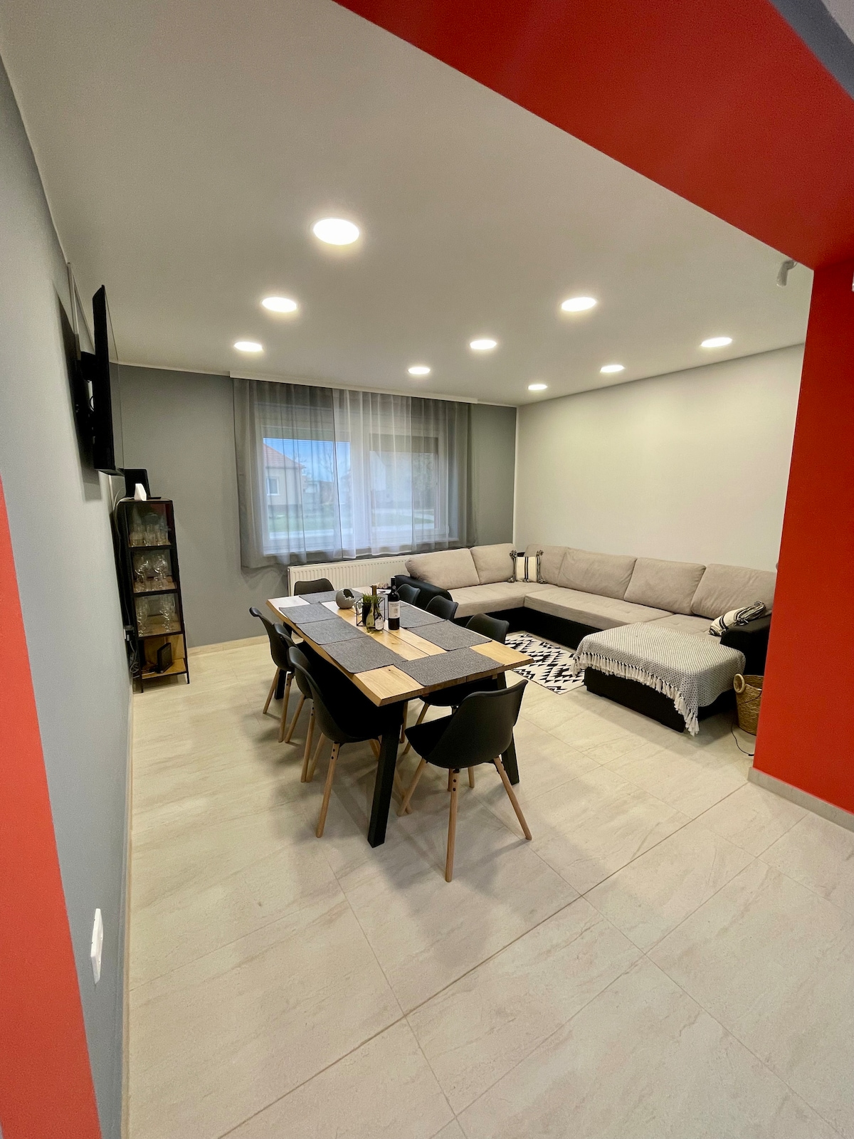A modern living space features a spacious corner sofa in neutral tones, complemented by a dining table surrounded by black chairs. Large windows allow natural light to fill the room, accented by sheer curtains. A subtle color palette enhances the comfortable environment.