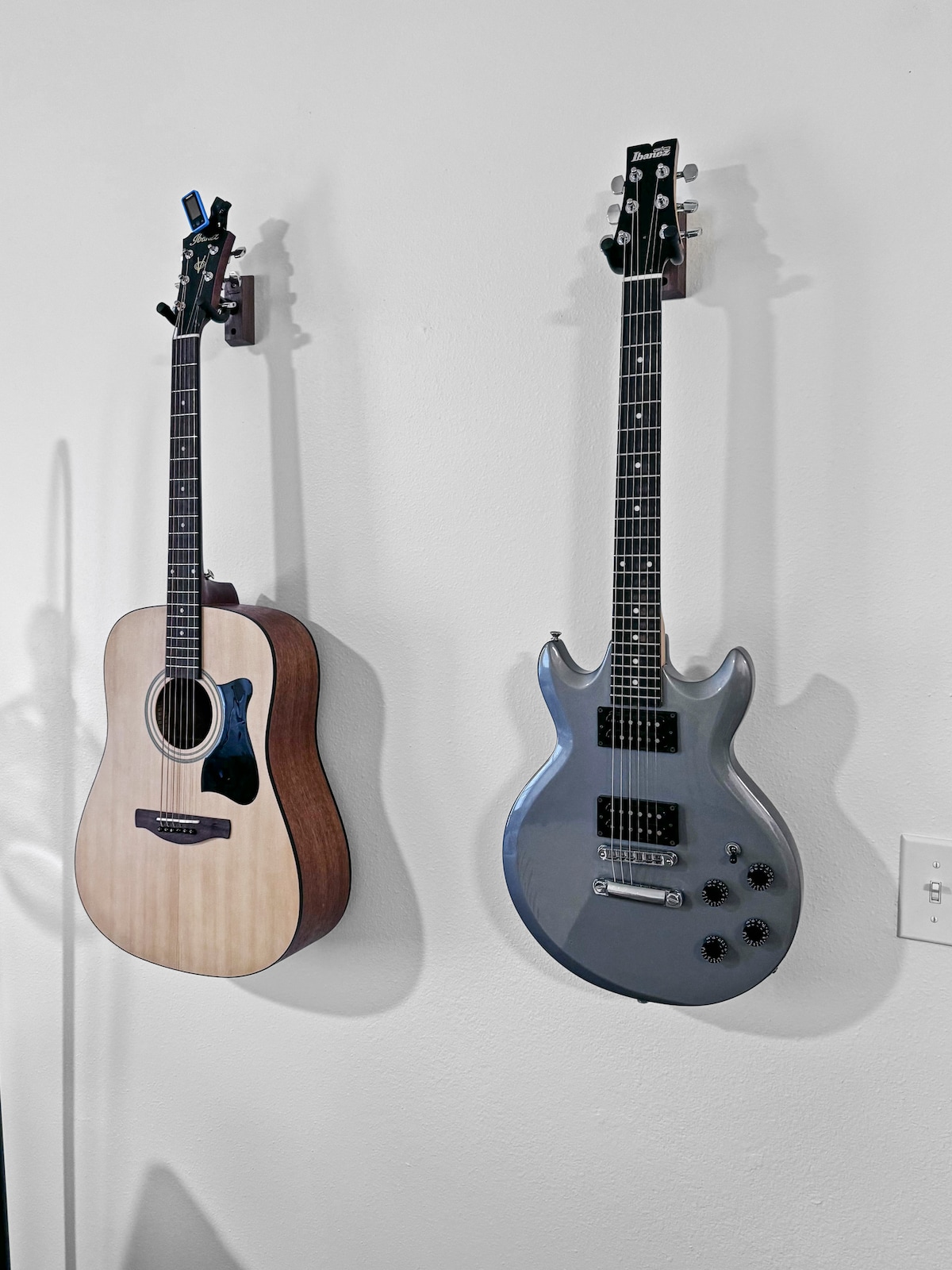 An acoustic guitar and an electric guitar are mounted on a light-colored wall. The acoustic guitar exhibits a natural wood finish, while the electric guitar features a sleek silver body with dark knobs. A power outlet is visible nearby.