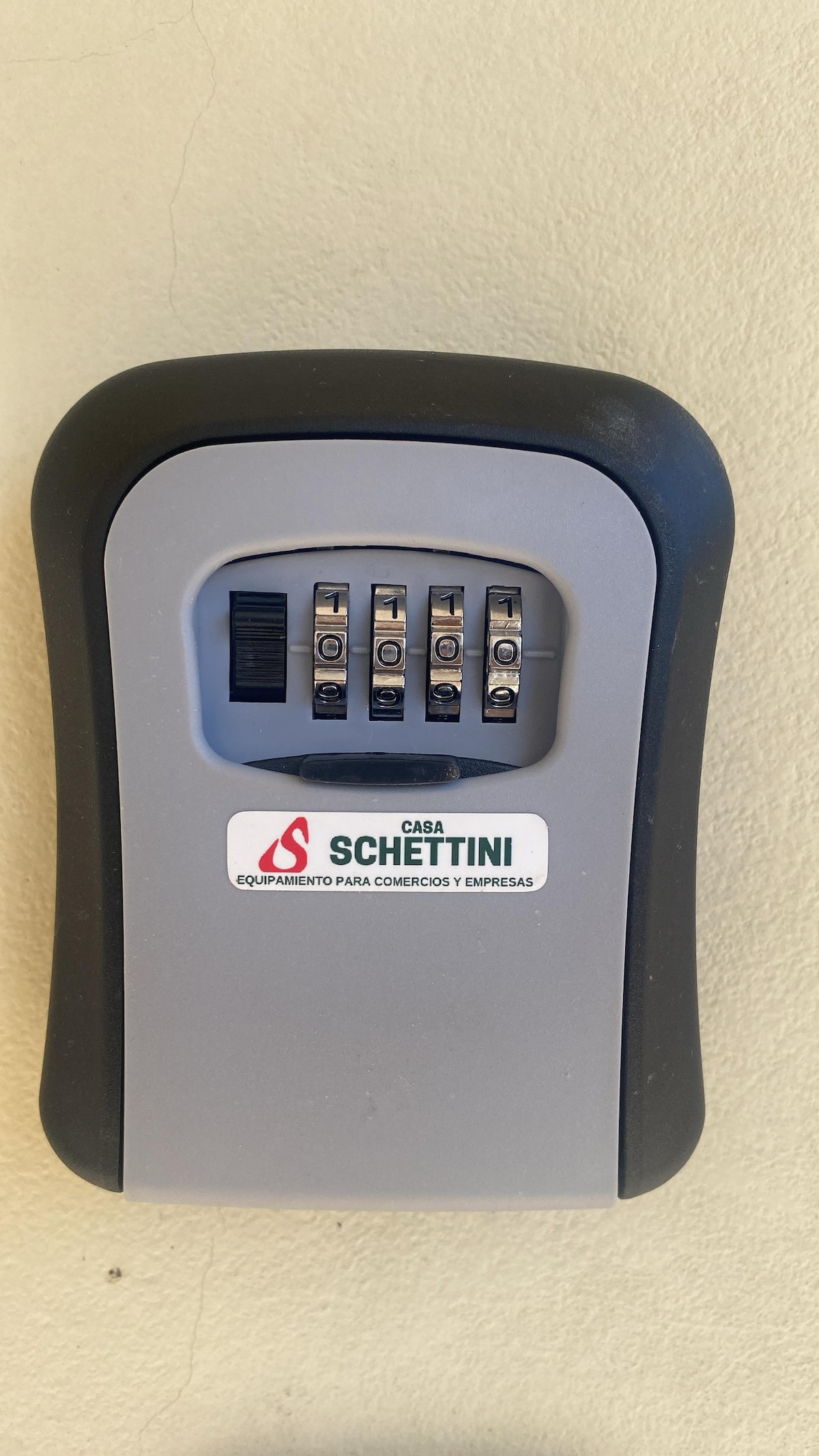 A secure key lockbox is displayed, featuring a combination dial with five numbered dials for code entry. The box is designed in gray and black, with a prominent logo labeled 'Casa Schettini' on the front. It is mounted on a smooth, neutral-colored surface.